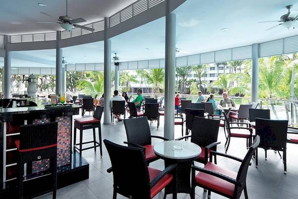Loungebereich des Riu Naiboa Hotels mit Tischen, Stühlen und Blick auf Palmen und Gebäude.