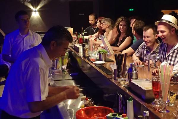 Ein Barkeeper mischt Getränke in einer Hotelbar mit blauer Beleuchtung, während Gäste an der Theke sitzen.