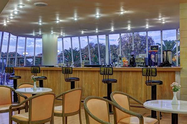 Innenansicht der Bar im Giannoulis Santa Marina Beach Hotel mit runden Tischen, Stühlen und einer Theke mit Getränken.