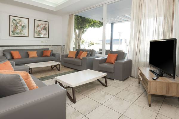 Wohnbereich des Kosta Palace City Hotels mit grauen Sofas, orangen Kissen und einer großen Fensterfront.