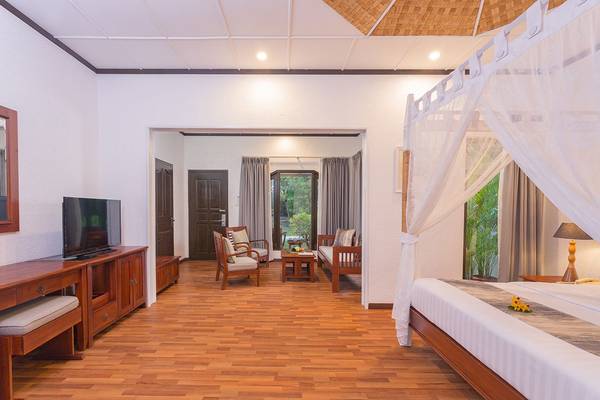 Innenansicht eines Zimmers im Bandos Maldives Hotel mit Holzboden, Bett, Fernseher und Wohnzimmerbereich.
