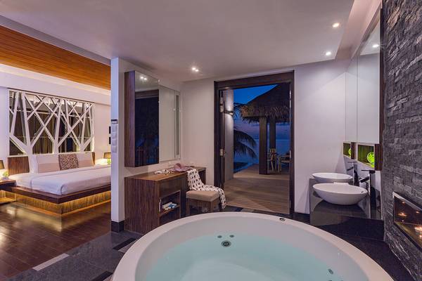 Innenansicht eines Zimmers im Bandos Maldives Hotel mit Badewanne, zwei Waschbecken, Kamin und Terrasse.