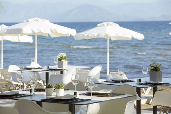 Strandrestaurant des Capo Di Corfu Hotels mit Tischen und Stühlen unter weißen Sonnenschirmen mit Blick auf das Meer.