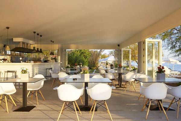 Innenbereich des Capo Di Corfu Hotels mit Esstischen und Stühlen, Blick auf eine Bar und den Außenbereich.