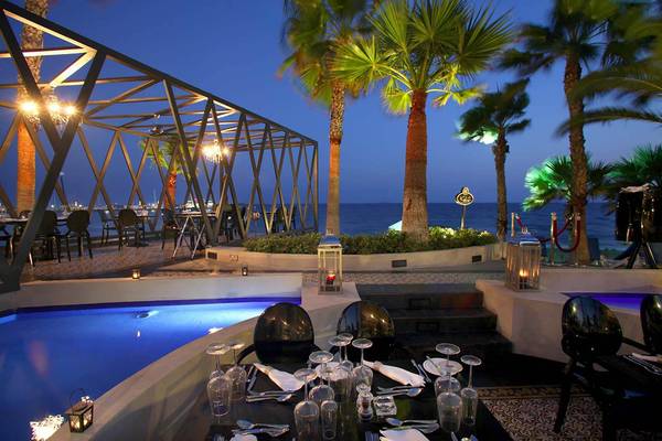 Außenbereich des GrandResort Limassol bei Nacht mit beleuchtetem Pool und Tischstühlen.