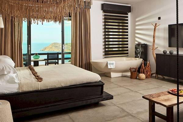 Myconian Utopia Resort Zimmer mit Meerblick, Bett, Badewanne, Fernseher, Tisch mit Getränken, Aussicht auf das Meer und Inseln.