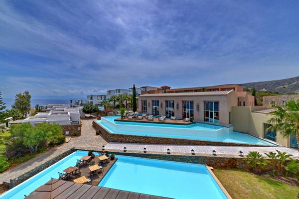 Luftaufnahme des Elounda Village Resort & Spa mit zwei Swimmingpools, Sonnenliegen und Bergpanorama.