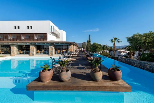 Außenpool im Elounda Village Resort & Spa mit Holzbrücke und Tischen, umgeben von Palmen und Gebäuden.