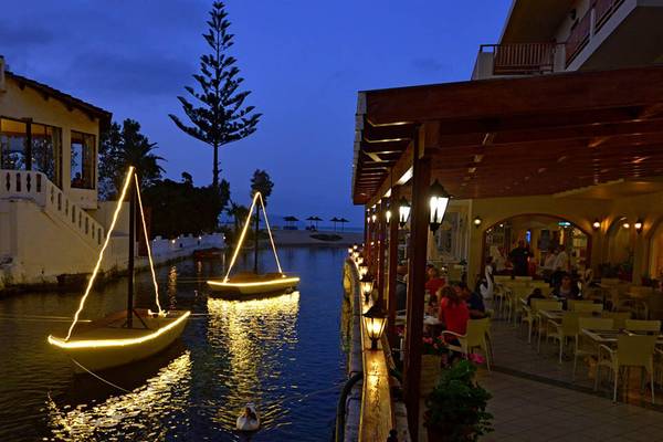 Abendansicht des Kalyves Beach Hotels mit beleuchteten Booten und Restaurant am Wasser.