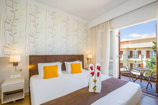 Zimmer im Kalyves Beach Hotel mit Balkon, Bett, Nachttischen und Ausblick auf eine Terrasse.