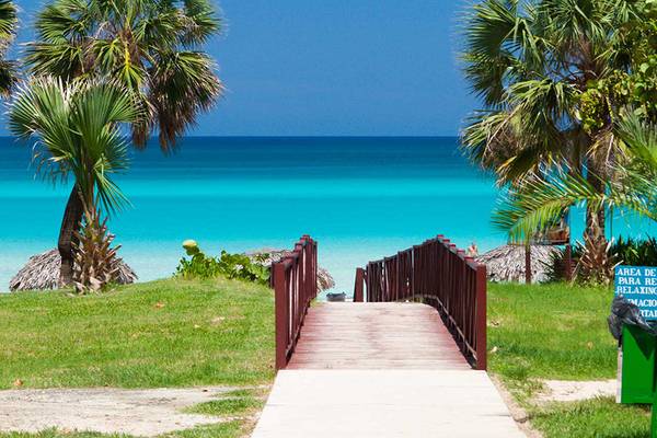 Eine Holzbrücke führt vom Hotel Occidental Arenas Blancas zum Strand mit türkisfarbenem Wasser.