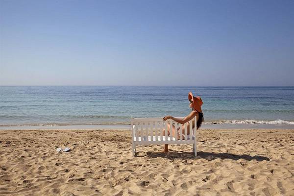 Frau in Sonnenhut sitzt auf einer Bank am Strand vor dem Alion Beach Hotel