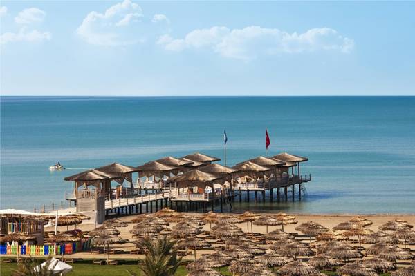 Strandansicht des Maxx Royal Belek Golf Resorts mit vielen Strohhütten und einem Boot im Wasser