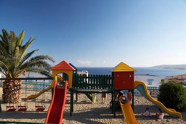 Spielplatz am Strand des Mitsis Family Village Beach Hotels mit Kindern auf den Spielgeräten und Blick auf das Meer.