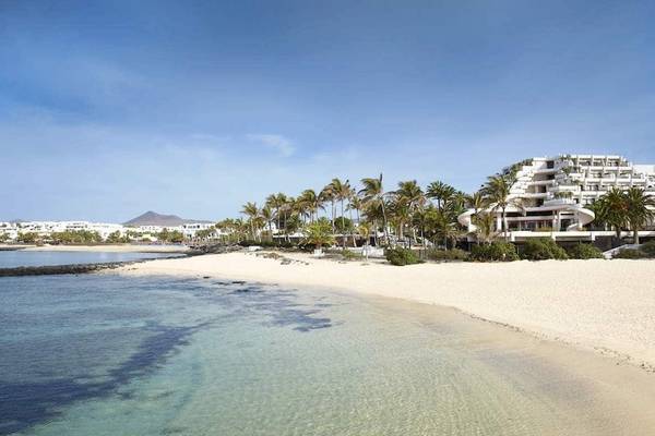 Außenansicht des Melia Salinas Hotels an einem weißen Sandstrand mit Palmen und Gebäuden im Hintergrund.