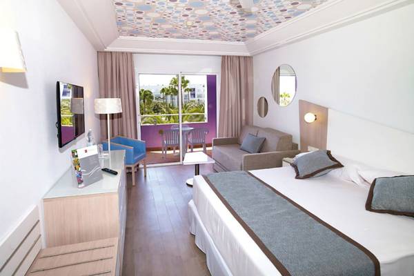 Innenansicht eines Hotelzimmers im Riu Gran Canaria mit Bett, Sofa, Fernseher und Balkon.