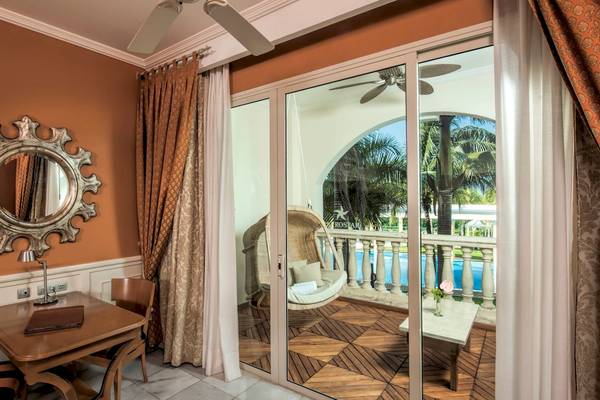 Innenansicht eines Zimmers im Iberostar Grand Paraiso mit Tisch, Stühlen, Spiegel und offenem Balkon mit Poolblick.