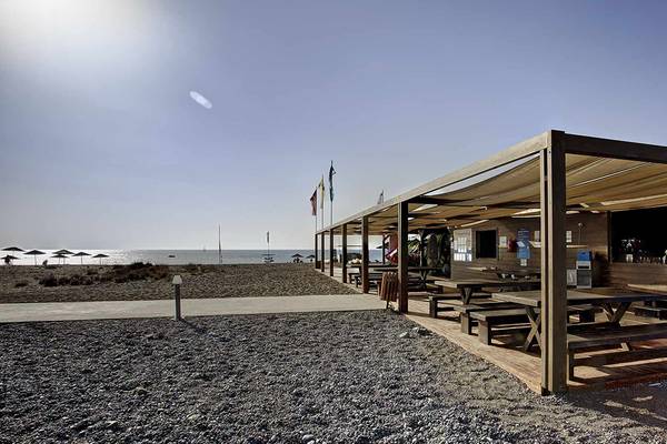 Außenbereich des Club Plimmiri Hotels mit Holztischen und Bänken unter einem Sonnendach am Strand.