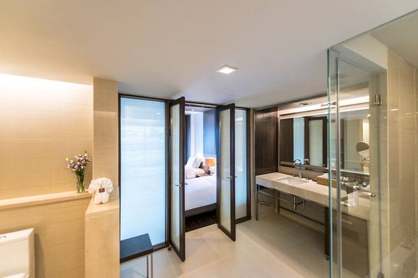 Badezimmer im Pathumwan Princess Hotel mit Glastür, Waschbecken und Bett im Hintergrund.
