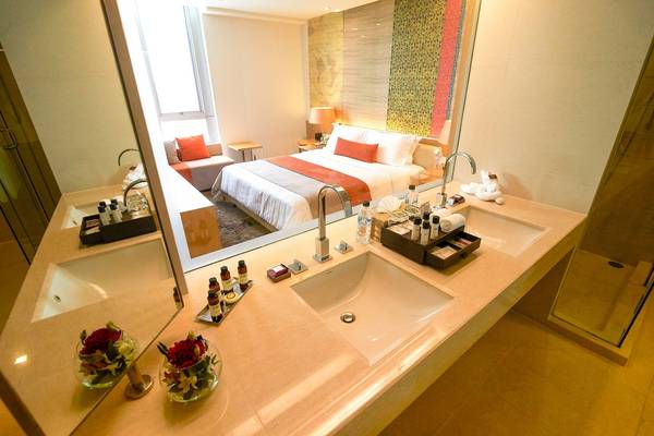 Badezimmer des Pathumwan Princess Hotels mit Waschbecken und Spiegel, dahinter ein Bett.