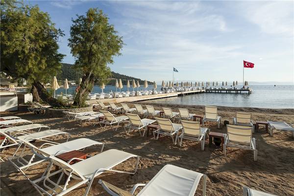 Strand mit Lounge-Sesseln und einem Steg, dahinter das Voyage Torba Hotel.