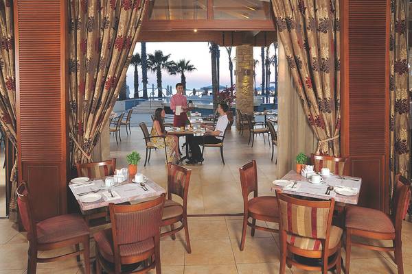 Innenansicht des Restaurants im Alexander The Great Hotel mit Gästen und Palmen im Hintergrund.