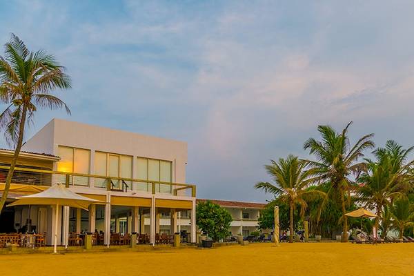Außenansicht des Jetwing Sea Hotels mit weißem Gebäude, Terrassen, Sonnenschirmen und Palmen am Strand