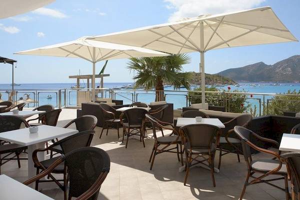 Terrasse des Universal Hotel Aquamarin mit Tischen und Stühlen unter großen Schirmen mit Blick auf das Meer.