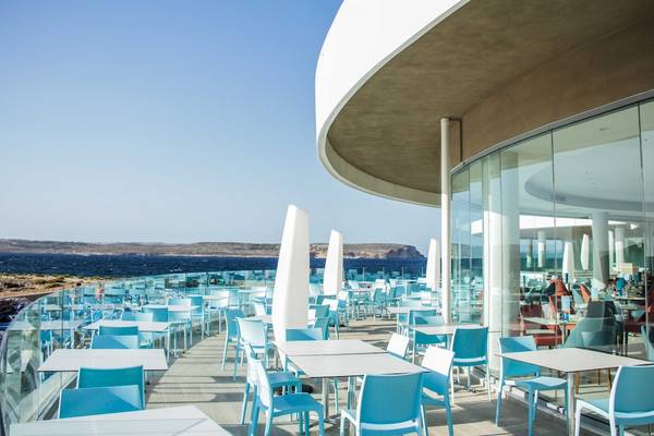 Terrasse des Ramla Bay Resorts mit Blick auf das Meer und blaue Tische und Stühle.