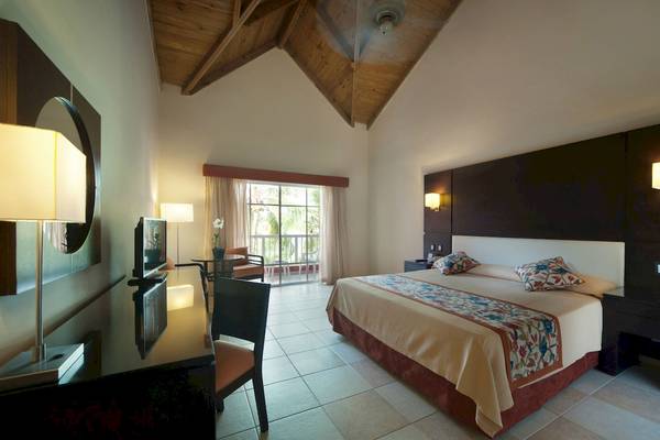 Innenansicht eines Hotelzimmers im Caribe Deluxe Princess Hotel mit Bett, Schreibtisch und Balkon.