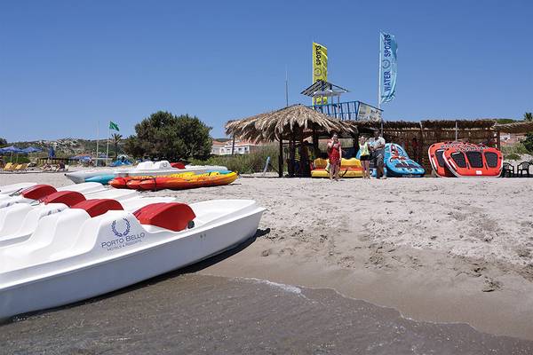 Strand des Porto Bello Hotels mit mehreren Booten und Gästen in der Nähe eines Sonnenschirms.
