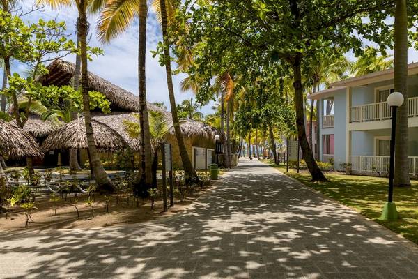 Der Eingang des Playabachata Spa Resorts mit Gehweg, Palmen und strohgedeckten Dächern