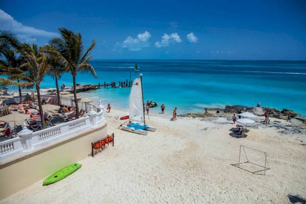 Strandszene des Riu Cancun Hotels mit weißen Sand, blauem Meer, Segelboot, Sonnenschirmen und Palmen