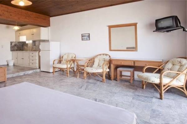 Ein modernes Schlafzimmer im Dedalos Beach Hotel mit weißem Bett, Holzmöbeln und einer Küche im Hintergrund.