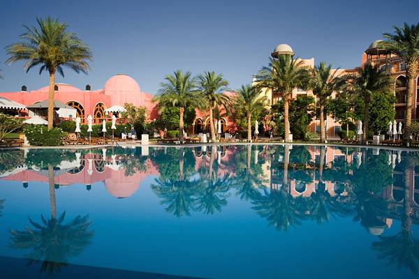 Ein Swimmingpool vor dem Grand Resort Hurghada Hotel mit spiegelnder Wasseroberfläche und umgeben von Palmen und Gebäuden.