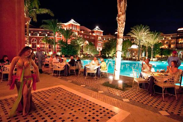 Eine Frau in einem farbenfrohen Kostüm tanzt neben einem Pool, während Gäste in einem Hotelrestaurant bei Nacht essen.