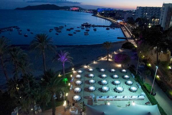Luftaufnahme von einer abendlichen Feier am Strand mit vielen gedeckten Tischen und einer Hotelanlage im Hintergrund.