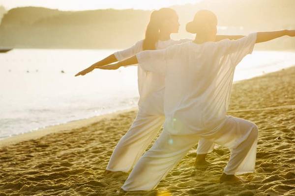 Zwei Personen praktizieren Yoga auf einem Strand bei Sonnenaufgang.