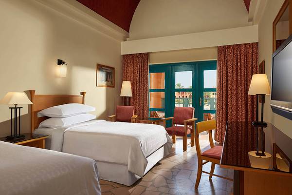 Doppelzimmer im Sheraton Miramar Resort El Gouna mit zwei Betten, Sessel, Schreibtisch und Fenstern.