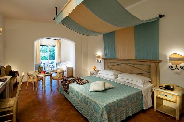 Interieur des Colonna Grand Hotel Capo Testa mit einem Bett, Tisch und Stühlen.
