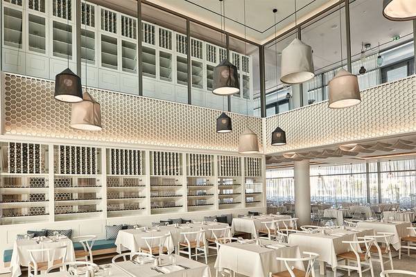 Innenansicht des Restaurants im Atlantica Dreams Resort mit weißen Tischen und Stühlen, dekorativen Lampen und einer Theke mit Weinflaschen.