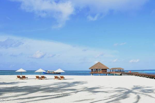 Strand des Noku Maldives mit weißen Sonnenschirmen, Liegestühlen, runden Hütten und einem Boot