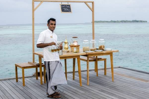 Mann, der vor einem Tisch am Strand des Noku Maldives steht und Getränke serviert.