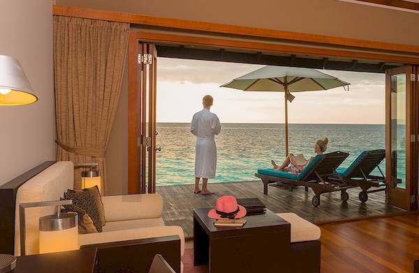 Innenansicht des Wohnzimmers im Veligandu Island Resort & Spa mit Meerblick, zwei Personen auf Sonnenliegen und einem offenen Glastürbereich.