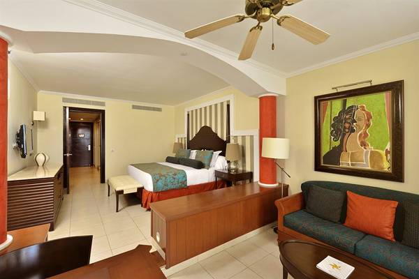 Innenansicht einer Suite im Iberostar Grand Rose Hall Hotel mit Bett, Sofa, Tisch und Gemälde.