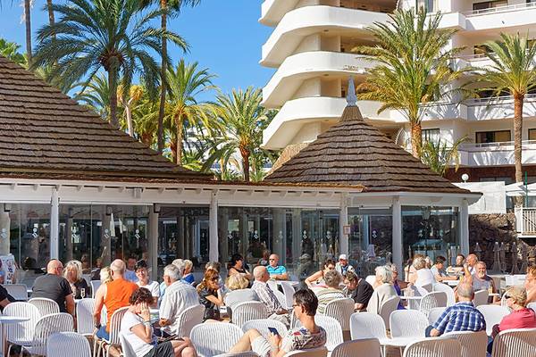Menschen sitzen auf der Terrasse des Servatur Waikiki Hotels vor Palmen und Gebäuden.