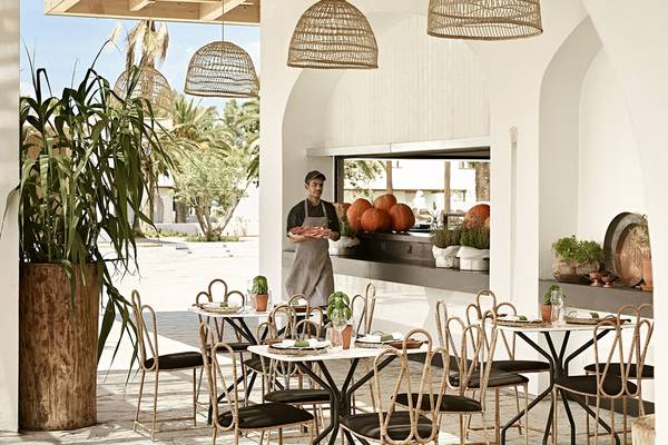 Ein Restauranttisch im Freien, mit dem Küchenchef, der vor einem Fenster steht, das auf einen Garten mit Kürbissen und Pflanzen hinausblickt.