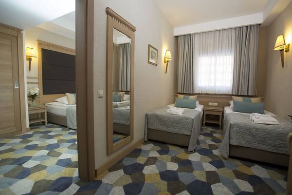 Innenansicht eines Doppelzimmers im Fame Residence Lara & Spa Hotel mit drei Betten, einem Spiegel und einem Tisch mit Blumen.