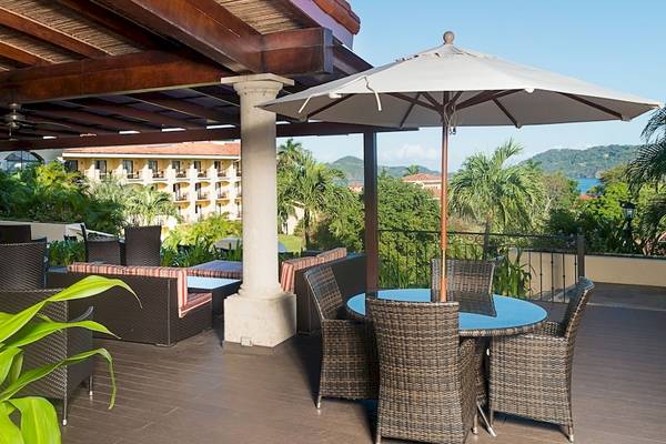 Terrasse des Occidental Papagayo Hotels mit Tischen, Stühlen und einem Schirm, Blick auf Gebäude und Landschaft