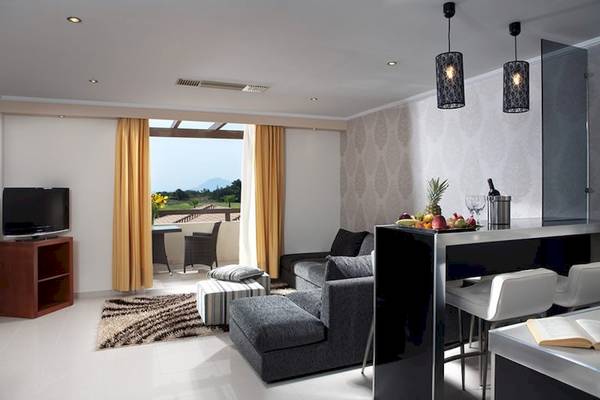 Wohnzimmer des Olympia Golden Beach Resorts mit Fernseher, Couch, Tisch und Balkonzugang.